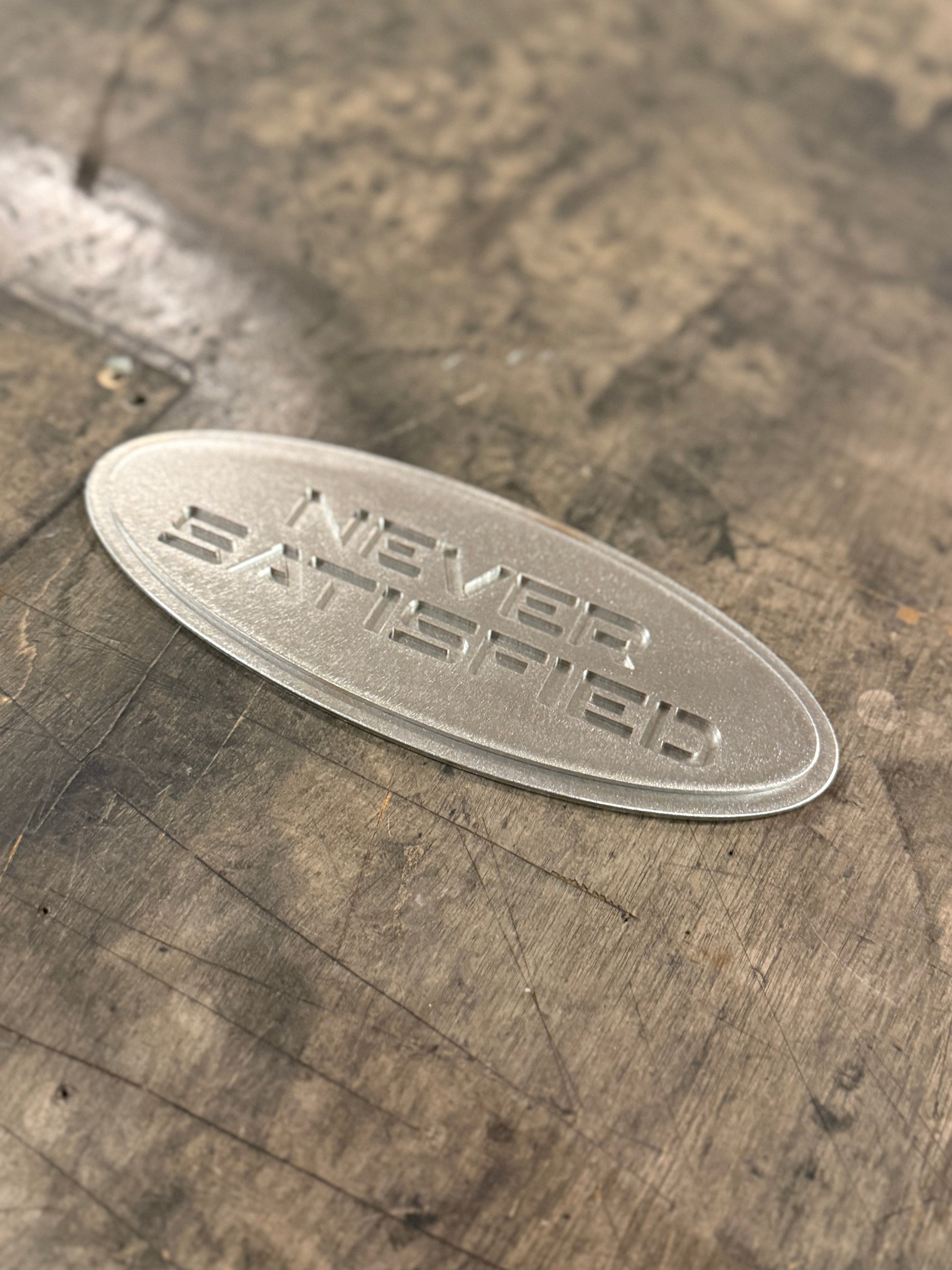 "Never Satisfied" Emblem (Ford 7" Universal Fitment) raw steel 